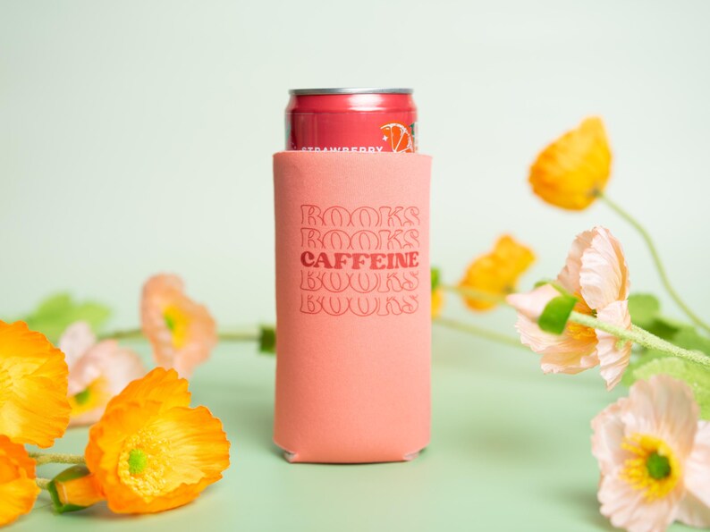 Op de afbeelding: Een koraalkleurige drankisolator met de woorden "BOOKS CAFFEINE BOOKS" in een retro lettertype. Een rood blikje aardbeiensoda zit in de isolator. De achtergrond toont oranje en roze bloemen op een lichtgroen oppervlak.