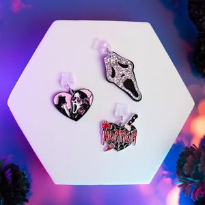 Op de afbeelding: Drie Halloween-oorbellen op een wit zeshoekig oppervlak. Eén is een sprankelend zilveren masker, een andere een zwart hart met een mes en het woord "HORROR", en de derde een roze hart met een eng gezicht.