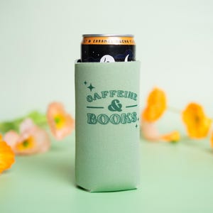 Op de afbeelding: Een lichtgroene blikjeskoeler met de tekst "Caffeine & Books" in turquoise. Een zwarte en gouden blikje zit erin. Oranje en roze bloemen zijn op de achtergrond.