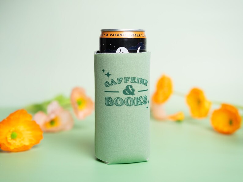 Op de afbeelding: Een lichtgroene blikjeskoeler met de tekst "Caffeine & Books" in turquoise. Een zwarte en gouden blikje zit erin. Oranje en roze bloemen zijn op de achtergrond.