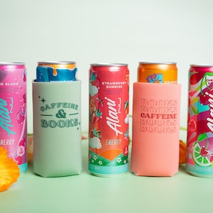 Op de afbeelding: Een assortiment Alani Nu energiedrankblikjes in verschillende smaken, waaronder Pink Slush en Strawberry Sunrise. De blikjes worden getoond met bijpassende drinkkoelers met de tekst "Caffeine & Books" en "Books Caffeine Books".