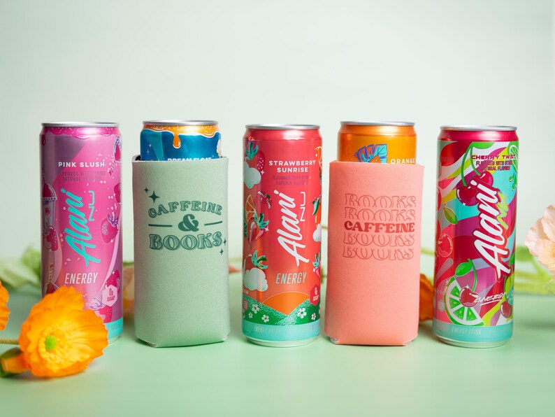 Op de afbeelding: Een assortiment Alani Nu energiedrankblikjes in verschillende smaken, waaronder Pink Slush en Strawberry Sunrise. De blikjes worden getoond met bijpassende drinkkoelers met de tekst "Caffeine & Books" en "Books Caffeine Books".