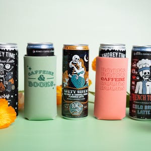 Op de afbeelding: Een assortiment koud gebrouwen koffieblikken met unieke ontwerpen en tekst. De blikken bevatten illustraties van een skelet, een zeemeermin en een chef-kok. Sommige blikken zitten in groene en koraalkleurige blikjeskoelers met de tekst "CAFFEINE & BOOKS" en "BOOKS CAFFEINE BOOKS".
