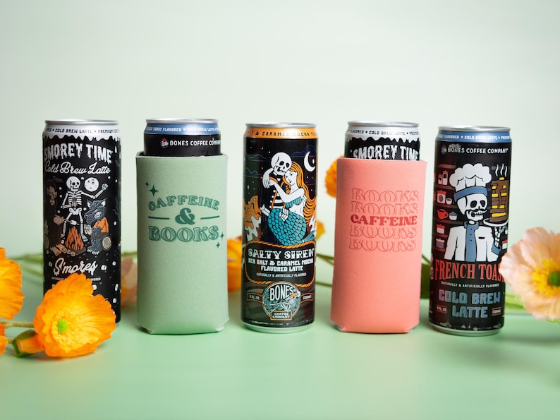 Op de afbeelding: Een assortiment koud gebrouwen koffieblikken met unieke ontwerpen en tekst. De blikken bevatten illustraties van een skelet, een zeemeermin en een chef-kok. Sommige blikken zitten in groene en koraalkleurige blikjeskoelers met de tekst "CAFFEINE & BOOKS" en "BOOKS CAFFEINE BOOKS".