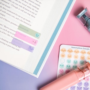 May include: An open book with three translucent adhesive page markers in light green, pink, and purple, each featuring a small emoji icon. A sheet of round annotation emoji stickers and two highlighter pens in muted blue and coral are placed nearby.