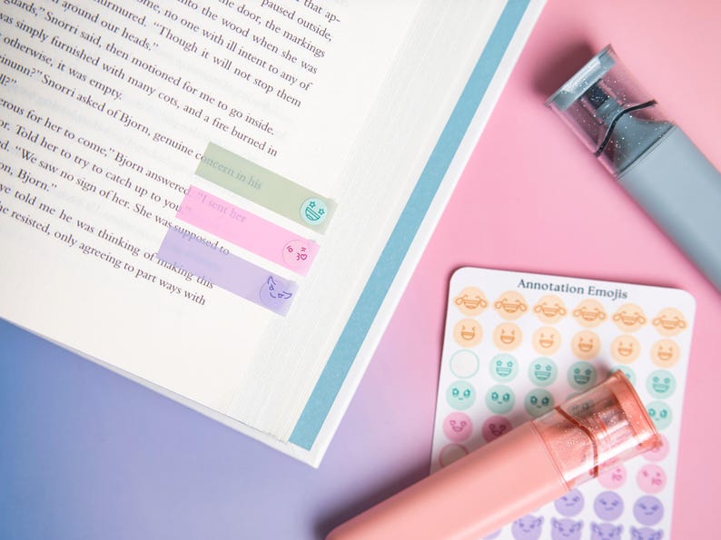 May include: An open book with three translucent adhesive page markers in light green, pink, and purple, each featuring a small emoji icon. A sheet of round annotation emoji stickers and two highlighter pens in muted blue and coral are placed nearby.