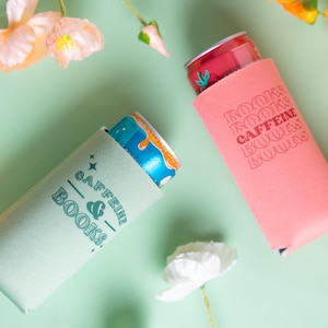 Op de afbeelding: Twee drankisolatoren, &eacute;&eacute;n lichtgroen en &eacute;&eacute;n roze, elk met een blikje. De lichtgroene isolator heeft de tekst "Caffeine & Books". De roze isolator heeft de tekst "Books Caffeine Books". Bloemenaccenten omringen de isolatoren op een lichtgroene achtergrond.