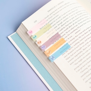May include: An open book with white pages and black printed text, featuring a series of colorful, translucent adhesive page markers attached to the edge of the paper. The markers are pink, yellow, peach, purple, orange, and blue, each with a small icon.