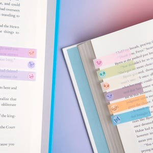 May include: A set of translucent, pastel-colored adhesive page markers attached to the edges of open book pages. Each rectangular marker features a small, simple printed facial expression, including smiling, frowning, and surprised faces in various colors.