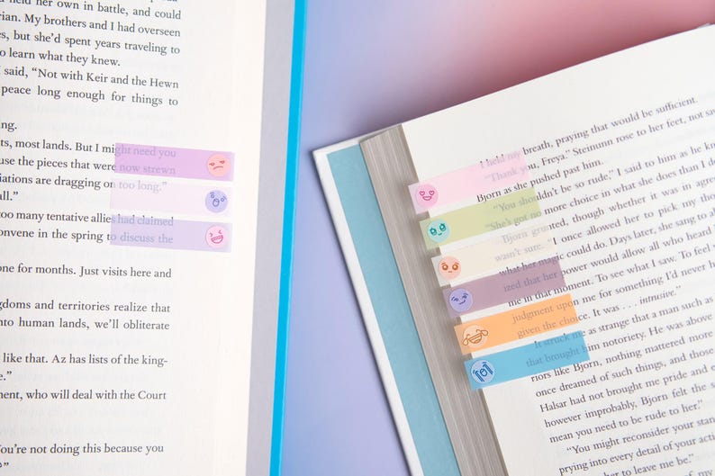 May include: A set of translucent, pastel-colored adhesive page markers attached to the edges of open book pages. Each rectangular marker features a small, simple printed facial expression, including smiling, frowning, and surprised faces in various colors.