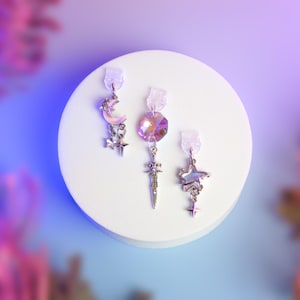 Peut inclure: Trois boucles d'oreilles sur le thème céleste sur un présentoir blanc. L'une présente un croissant de lune et des étoiles, une autre un cristal avec un charme d'épée, et la troisième une étoile et une petite étoile. Les boucles d'oreilles sont argentées.