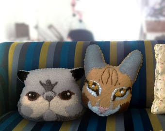 May include: Three plush animal pillows on a striped couch. The pillows are shaped like a cat, a raccoon, and a fox. The cat pillow is brown and white, the raccoon pillow is gray, and the fox pillow is orange and white.