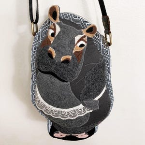 May include: A gray hippo-shaped crossbody bag with a black strap and gold-colored hardware. The bag features a white lace collar and a patterned background. The text "Wing's World" and "Dream Work Series" are visible.