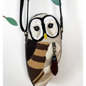 May include: A brown and tan owl-shaped crossbody bag with a brown leather feather charm. The bag has a black strap and is made of felt. The text "Wing's World Dream Work Series" is printed below the bag.