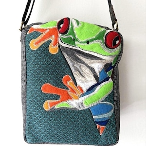 May include: A teal crossbody bag featuring an appliqué of a red-eyed tree frog. The frog has bright green, orange, and blue details. The bag has a black strap and the text "Wing's World" at the bottom.
