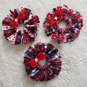May include: Three handmade fabric wreath ornaments. Each wreath features a circular design with red, white, and blue plaid and star-patterned fabric. A red bow and small white flower accent the center of each wreath. White string is attached for hanging.