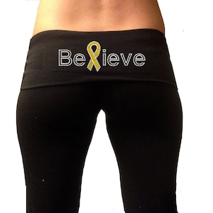 Könnte beinhalten: Schwarze Yogahose mit einem breiten Bund, der mit einem gelben Band und dem Wort "Believe" in weißen Strasssteinen verziert ist.