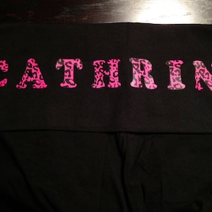 Op de afbeelding: Zwarte leggings met een roze luipaardprint met de tekst "CATHRINE" over de tailleband.