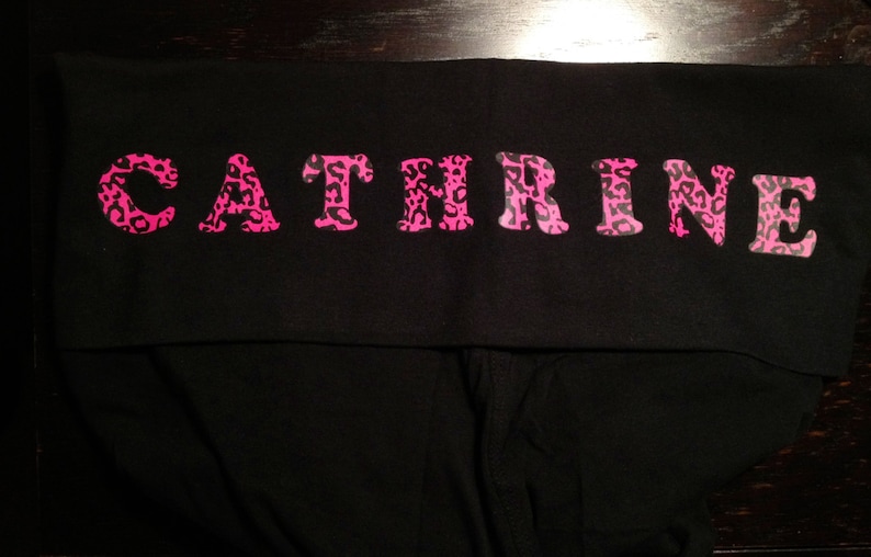 Op de afbeelding: Zwarte leggings met een roze luipaardprint met de tekst "CATHRINE" over de tailleband.