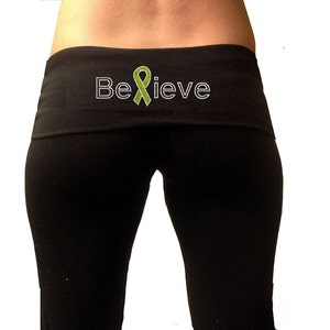 Op de afbeelding: Zwarte yogabroek met een brede tailleband met een groen lint en het woord "Believe" in witte strass steentjes.