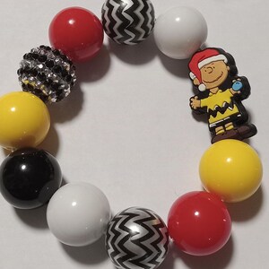 May include: A beaded bracelet featuring a variety of colorful beads. The bracelet includes red, white, black, and yellow beads, along with beads with a black and white zigzag pattern. A charm of Charlie Brown in a Santa hat is also included.