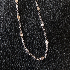May include: A delicate silver bracelet with small, round, clear gemstones evenly spaced along the chain. The bracelet has a lobster clasp closure and a small extension chain. The background is a black, textured surface.