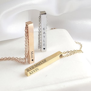 May include: Three personalized bar necklaces in rose gold, silver, and gold. Each pendant is engraved with names and dates. The necklaces hang on delicate chains. The background is a white surface with soft, white fabric.