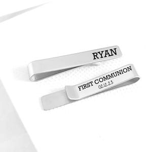 May include: Two silver-toned metal tie clips. One clip is engraved with the name RYAN in black capital letters. The second clip is engraved with the text FIRST COMMUNION and the date 02.12.23 in black capital letters and numbers.