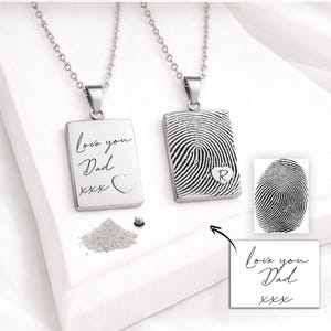 Op de afbeelding: Twee zilveren rechthoekige hangerkettingen. Eén hanger heeft de inscriptie "Love you Dad" met een hart en "xxx". De andere hanger heeft een vingerafdrukontwerp met de letter "R". Beide hangen aan delicate kettingen.