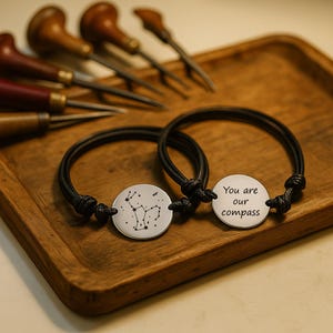 Può includere: Due braccialetti in corda nera con ciondoli circolari in metallo color argento. Un ciondolo mostra un motivo a costellazione nero, mentre l'altro è inciso con il testo "You are our compass" in un carattere semplice, scuro e senza grazie.