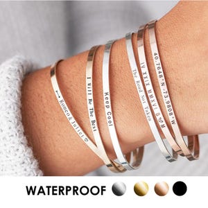 Könnte beinhalten: Eine Sammlung dünner, gravierter Metallarmbänder in Silber-, Gold- und Roségoldtönen. Jedes Armband hat eine andere Inschrift, darunter Sätze und Koordinaten. Das Wort "WATERPROOF" wird unter den Armbändern angezeigt.