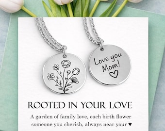 May include: Two silver-toned circular pendant necklaces on a white display card. One pendant features a black line-art floral bouquet, while the other is engraved with the text 'Love you Mom!' and a heart. The card reads 'ROOTED IN YOUR LOVE'.
