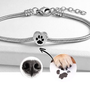 Könnte beinhalten: Ein silberfarbenes Schlangenkettenarmband mit einem herzförmigen Anhänger, der mit einem schwarzen Pfotenabdruck graviert ist. Das Armband verfügt über einen Karabinerverschluss und eine verstellbare Verlängerungskette. Zwei runde Einsätze zeigen eine Nase und einen Pfotenabdruck.