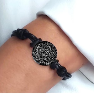 Könnte beinhalten: Ein schwarzes Kordelarmband mit einem runden Anhänger, der ein weißes Sternenkarten-Design zeigt. Der Anhänger präsentiert ein detailliertes Raster von Sternbildern auf schwarzem Grund und ist beidseitig mit Knoten befestigt.