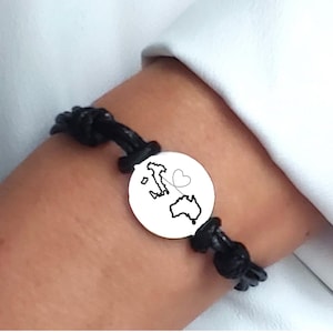 May include: A black cord bracelet featuring a circular silver-toned charm. The charm displays a black outline map of Italy and Australia connected by a dashed line that forms a heart shape in the center between the two landmasses.
