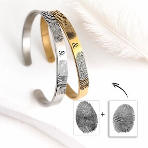 Könnte beinhalten: Zwei offene Armreifen, einer in Silber- und einer in Goldoptik, jeweils graviert mit einem einzigartigen schwarzen Fingerabdruckmuster und einem Et-Zeichen. Unter den Armreifen befinden sich zwei separate schwarz-weiße Fingerabdruckbilder mit einem Pluszeichen.