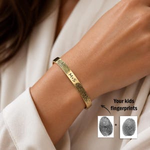 May include: A gold-toned metal cuff bracelet featuring two engraved black fingerprint patterns and the text M+S. Below the bracelet, a small graphic shows two distinct black fingerprint images with a plus sign and the text Your kids fingerprints.