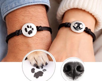 May include: Two black cord bracelets featuring circular silver-toned charms. One charm displays a black ink paw print, while the other charm displays a black ink nose print. The design is inspired by the paw and nose prints shown in circular insets.
