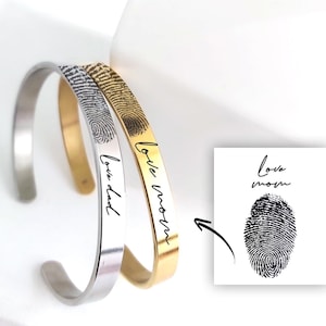 Può includere: Due bracciali rigidi in metallo, uno color argento e uno color oro, incisi con un'impronta digitale nera e il testo in corsivo "love dad" o "love mom". Un grafico quadrato mostra il testo "love mom" sopra un'impronta digitale con una freccia.