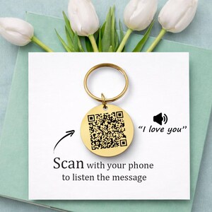 Könnte beinhalten: Goldfarbener Schlüsselanhänger mit rundem Anhänger mit QR-Code. Der Text "Scan with your phone to listen the message" und "I love you" sind ebenfalls abgebildet. Der Schlüsselanhänger befindet sich auf einer weißen Karte mit weißen Tulpen.