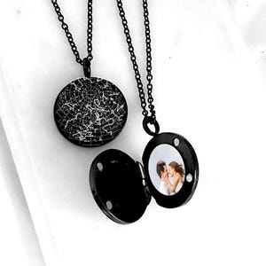 Puede incluir: Dos collares con relicario de metal negro y cadenas finas. Un relicario está cerrado y muestra un mapa estelar blanco sobre un fondo circular negro. El otro relicario está abierto, revelando una pequeña fotografía circular de dos personas.
