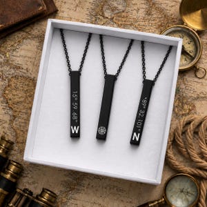 May include: Three black vertical bar pendants on matching black chains, presented in a white box. The left pendant features the text 15 59.68 and W, the center has a compass rose icon, and the right displays 89 32.101 and N in white lettering.