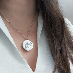May include: A silver necklace with a round pendant featuring a floral design. The pendant has a white background with black line drawings of various flowers. The necklace is worn against a white shirt.