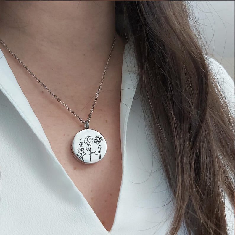 May include: A silver necklace with a round pendant featuring a floral design. The pendant has a white background with black line drawings of various flowers. The necklace is worn against a white shirt.