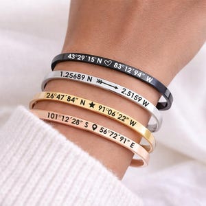 May include: Four metal bangles in black, silver, gold, and rose gold are stacked on a wrist. Each bangle has engraved coordinates and symbols. The black bangle reads "43°29'15" N 83°12'94" W".