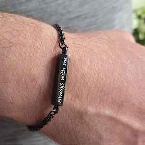 Könnte beinhalten: Ein schwarzes Metallkettenarmband mit einem rechteckigen Stabanhänger. Der Stab ist mit dem weißen Text Always with me in Schreibschrift graviert. Das Armband besteht aus einem Gliederkettendesign, das mit dem zentralen Stabelement verbunden ist.