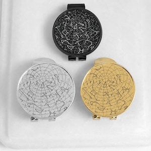 Peut inclure: Trois pinces à billets circulaires arborant un motif de carte céleste. Une pince est noir mat avec des constellations blanches, une est en argent poli avec des constellations noires, et une est en or poli avec des constellations noires.