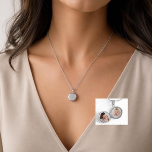 May include: A silver-coloured circular locket pendant on a delicate matching chain. The locket is shown closed in the main view and open in the inset, revealing two internal circular frames containing small photographs of a person and a baby.