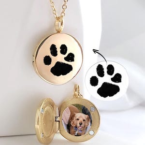 May include: A gold-toned circular locket pendant on a matching chain. The front of the locket features a black paw print design. The locket is shown open, revealing a circular photograph of a light brown dog inside the metal casing.