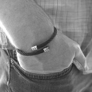 May include: Black and silver bracelet. The bracelet is made of a twisted, dark-coloured material and features silver decorative clasps at each end. The bracelet is worn on the wrist. The image is in black and white.